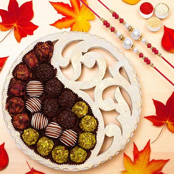 Round ornate tray half-filled with assorted chocolates and truffles on a light wooden table; autumn red and orange leaves and decorative beaded skewers surround the scene.